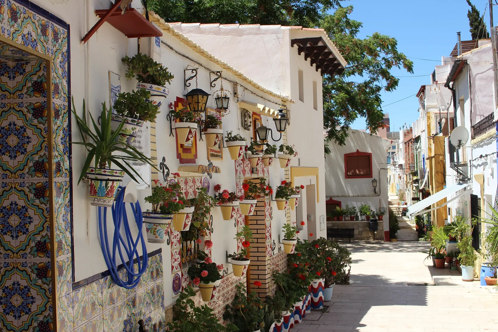 A sunlit lane in the Santa Cruz quarter of Alicante, its white walls covered in painted tiles, flower pots and lanterns; a general photograph of the quarter, not of the Marbeuf Palace